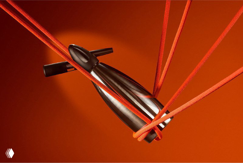 Металлический предмет и коричнево-красные верёвки в студийной композиции на оранжевом фоне рядом с элементами фирменной айдентики Gaya Athens и деталями полиграфии.