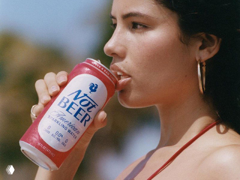 Женщина пьёт из банки Not Beer на размытом летнем фоне; красно‑белая упаковка стилизована под пиво, подчёркивается лайфстайл‑фотография.