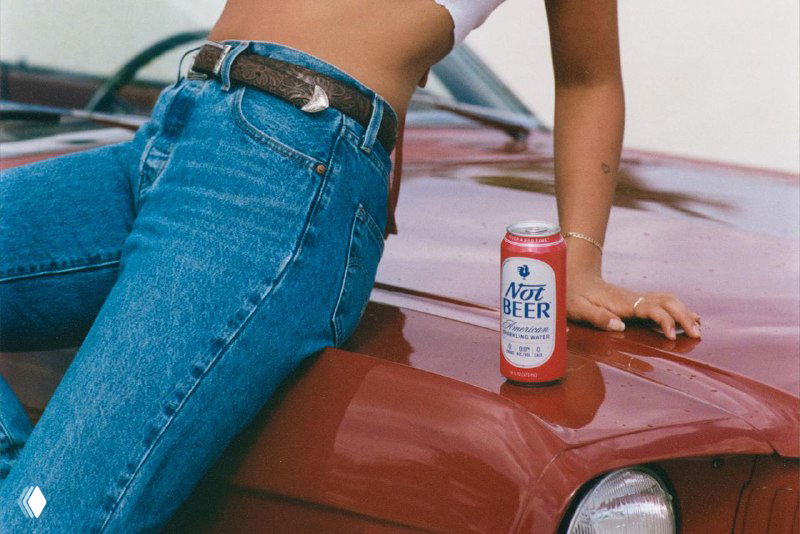 Банка Not Beer стоит на капоте ретро‑автомобиля рядом с ногами человека в джинсах; кадр подчёркивает винтажный стиль и упаковочный дизайн.