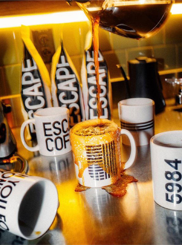 Крупный план кухонной сцены: кружки и баннеры Escape, кофе льётся в чашку — видны логотипы и предметы с брендингом кампании.