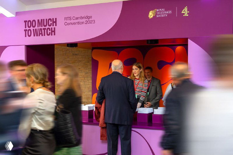 Зона регистрации конференции с баннером и большим цветным фоном, на котором размещена крупная вариативная типографика «Too Much To Watch», посетители в движении.