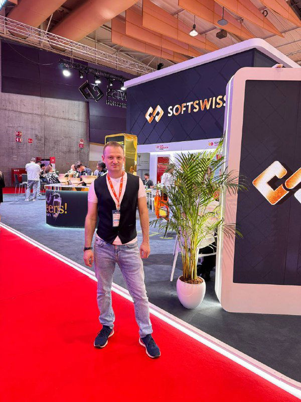 Representative standing on the red carpet by the SoftSwiss booth at SBC Summit Lisbon, wearing an event badge and casual outfit.