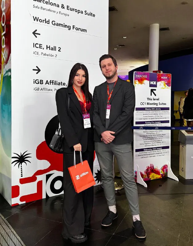 Two Zillion Games representatives posing in front of ICE event signage, one holding an orange Zillion-branded bag while both wear attendee lanyards and badges.