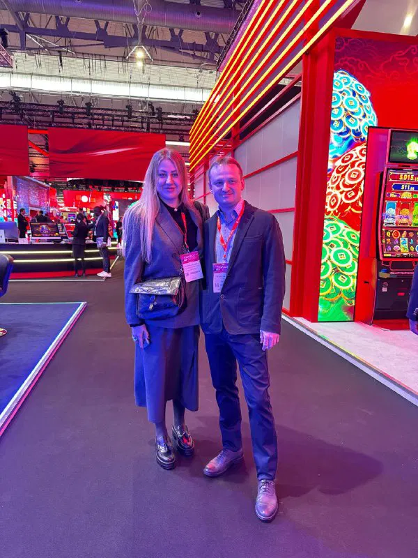Two attendees from Zillion Games posing on the exhibition floor with illuminated slot machine booths and colorful LED displays visible in the background.