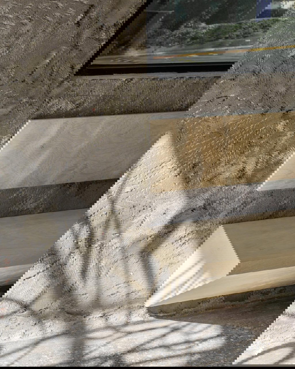 Фрагмент грубой штукатурки и деревянная лавка в интерьере: контраст текстур, простые формы и натуральное дерево в минималистичном пространстве.