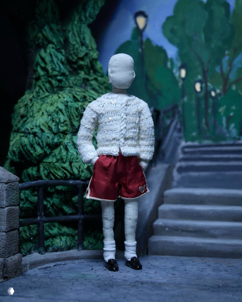 Кукла в белой вязаной кофте и коротких бордовых шортах на фоне миниатюрной лестницы и зелёных растительных декораций.