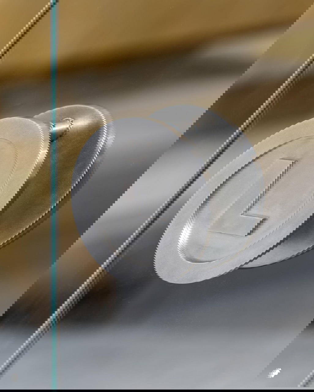 Крупный план металлической круглой дверной ручки с тиснёным логотипом «L» — элемент айдентики, выполненный в минималистичном промышленном стиле.