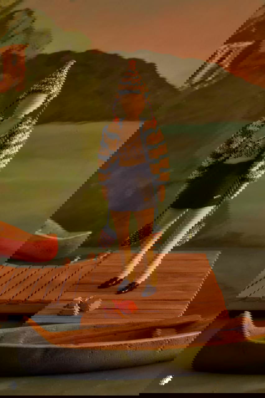 Миниатюрная кукла в полосатом вязаном комплекте с шапочкой стоит на пирсе рядом с каноэ — пастельные закатные цвета в кадре.