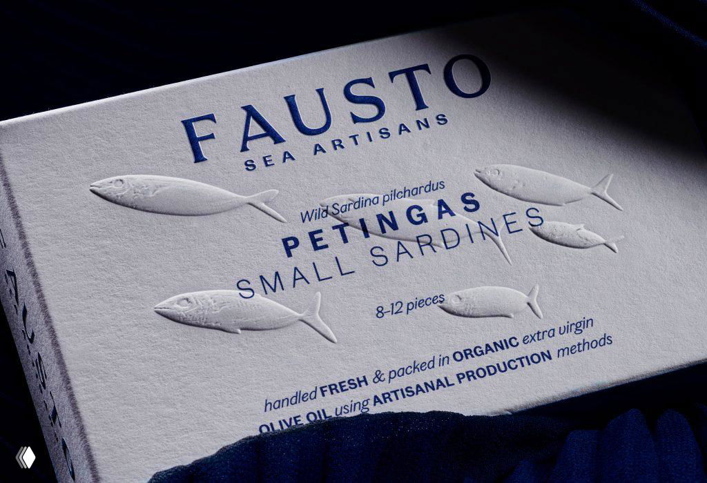 Крупный план тиснения на картоне: рельефные изображение рыб и надпись FAUSTO, видна фактура бумаги и глубина рельефа в естественном свете.