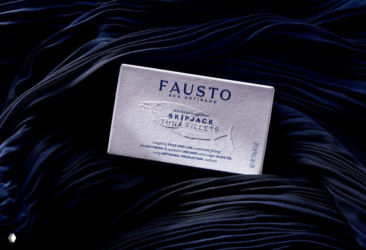 Вариант упаковки консервов FAUSTO для тунца с крупным логотипом и тиснением, расположенный на волнообразной тёмной ткани, подчёркивающей морскую тему бренда.