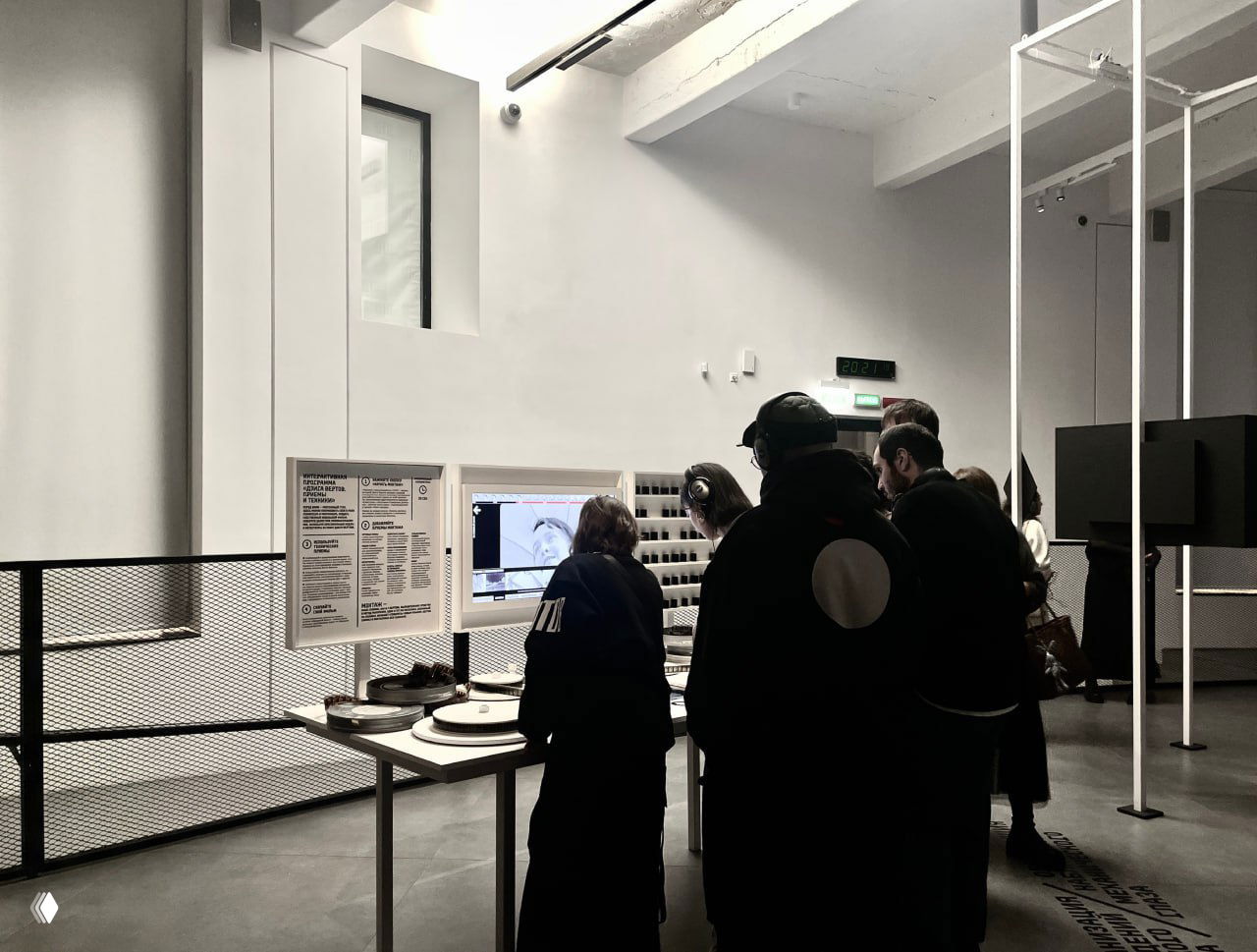 Посетители выставки у монтажного стола: люди с наушниками и экранами в минималистичном белом пространстве галереи, изучают интерфейс монтажа