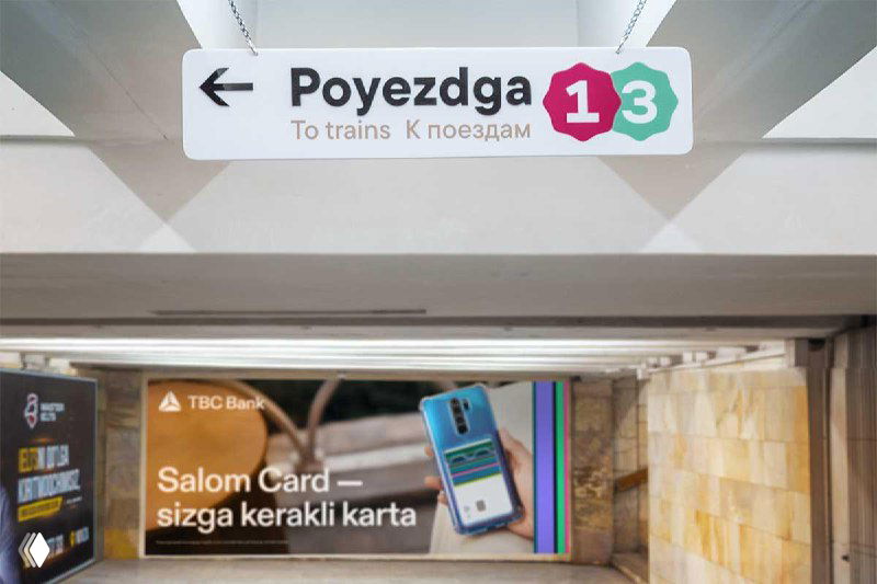 Крупный план подвесного указателя с надписью «Poyezdga», стрелкой и цветными значками линий на светлом фоне и размытым рекламным щитом позади.