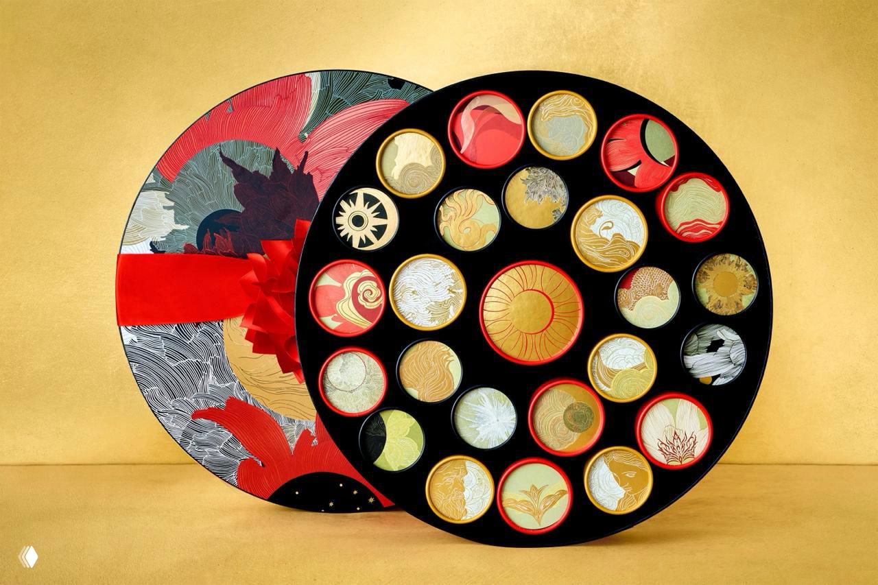 Вид сверху на раскрытую круглую коробку: аккуратно размещённые круглые ячейки с цветными и золотыми художественными крышками и ярким фоном.