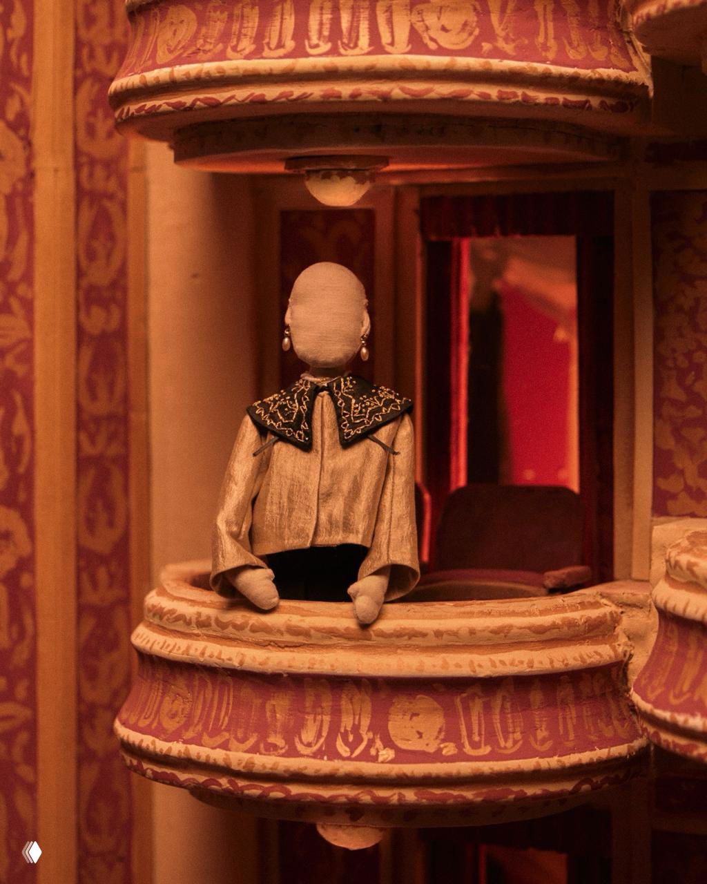 Кукла в светлом жакете с крупным воротником сидит в миниатюрном театральном ложу — интерьер тёплых тонов и театральная деталь.
