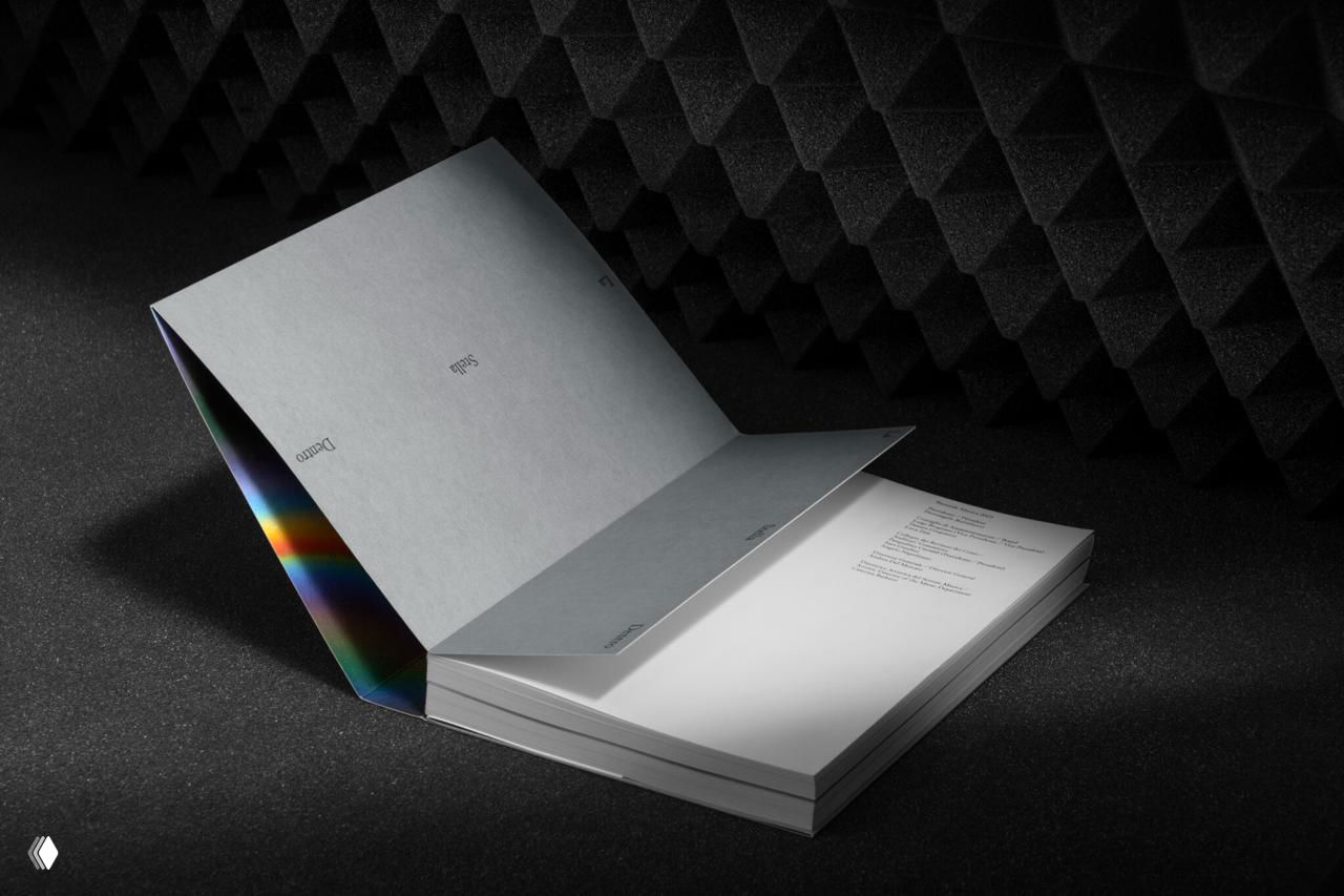 Каталог в раскрытом положении на текстурном тёмном фоне, видна внутренняя радужная вкладка и аккуратный шрифт на серой обложке.