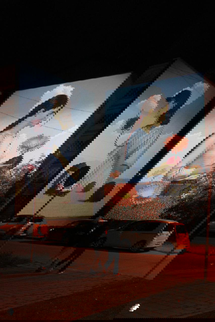 Ночной кадр: две панели триптиха подсвечены на фасадах домов, пешеходы проходят мимо, видна композиция из облаков, фигур и цветов на стенах.