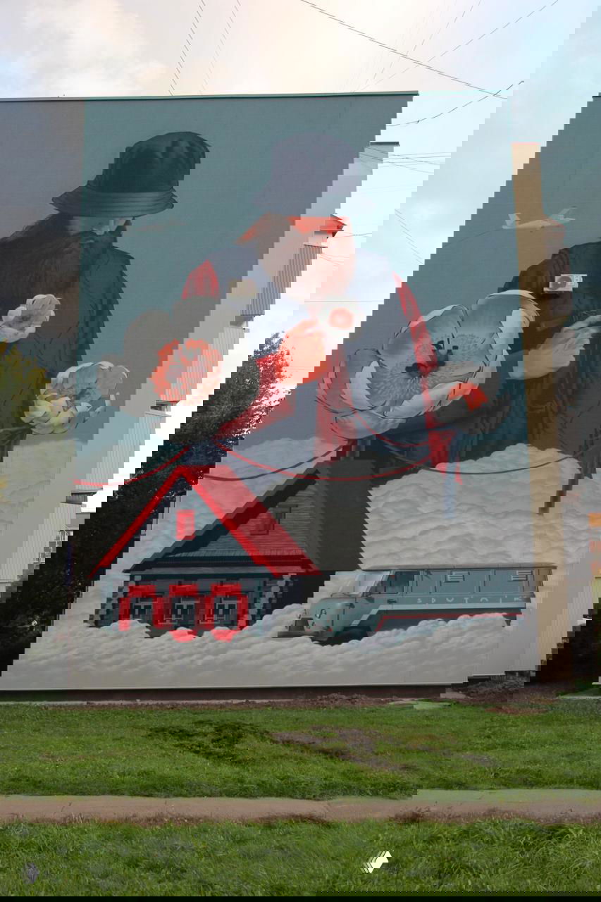 Мурал с большим мужчиной в панаме над домом и почтовой машиной, цветы и облака образуют мягкую «почву» для домика внизу фасада.