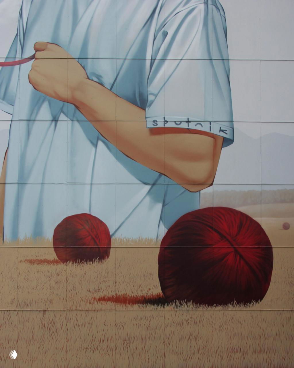 Деталь мурала: рука и часть футболки с подписью «sputnik», на переднем плане два больших клубка нитей на травяном поле.