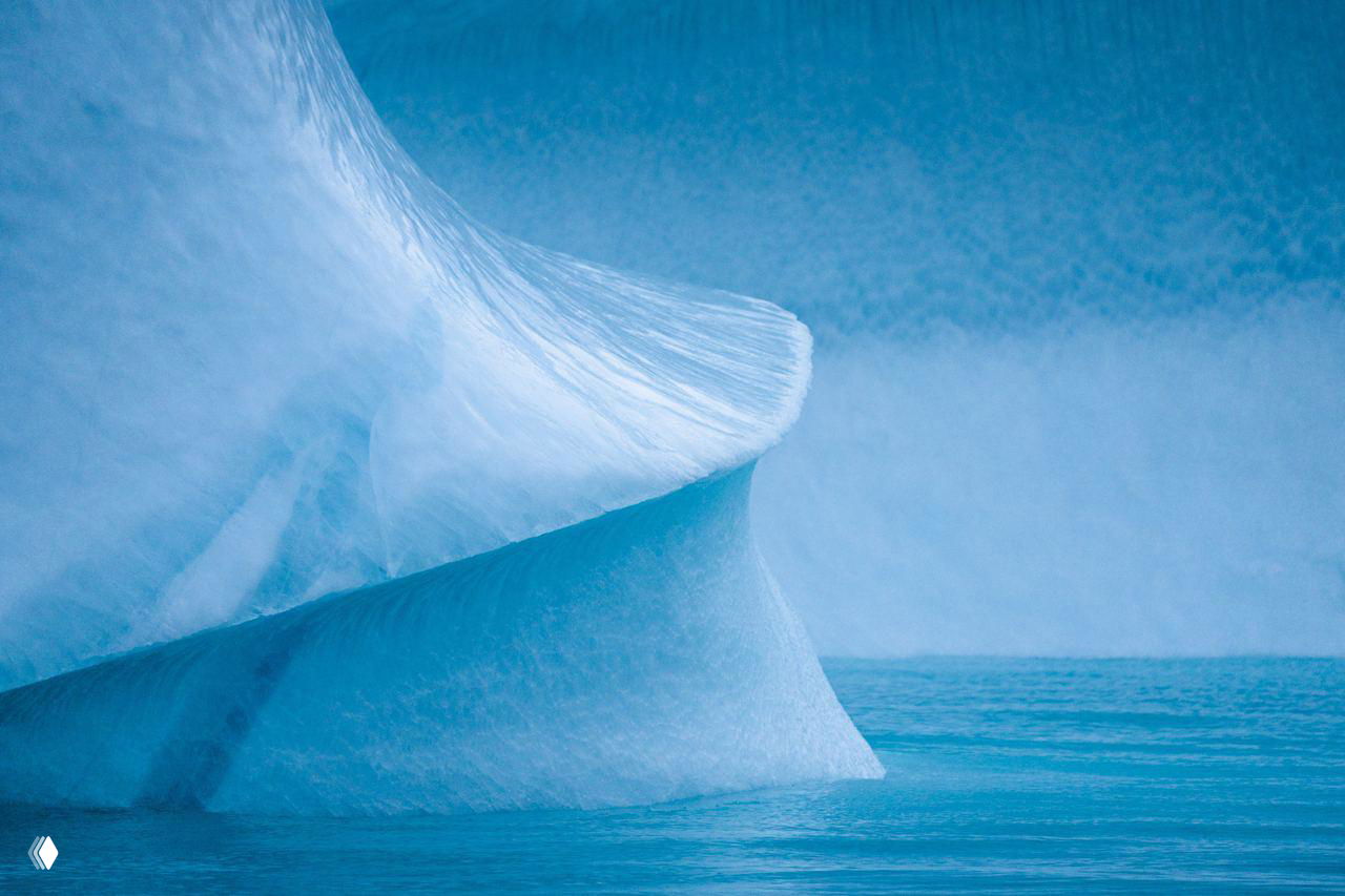 Крупный план ледяной кромки айсберга с выразительной текстурой и переливами синевы, снятый близко к поверхности воды.