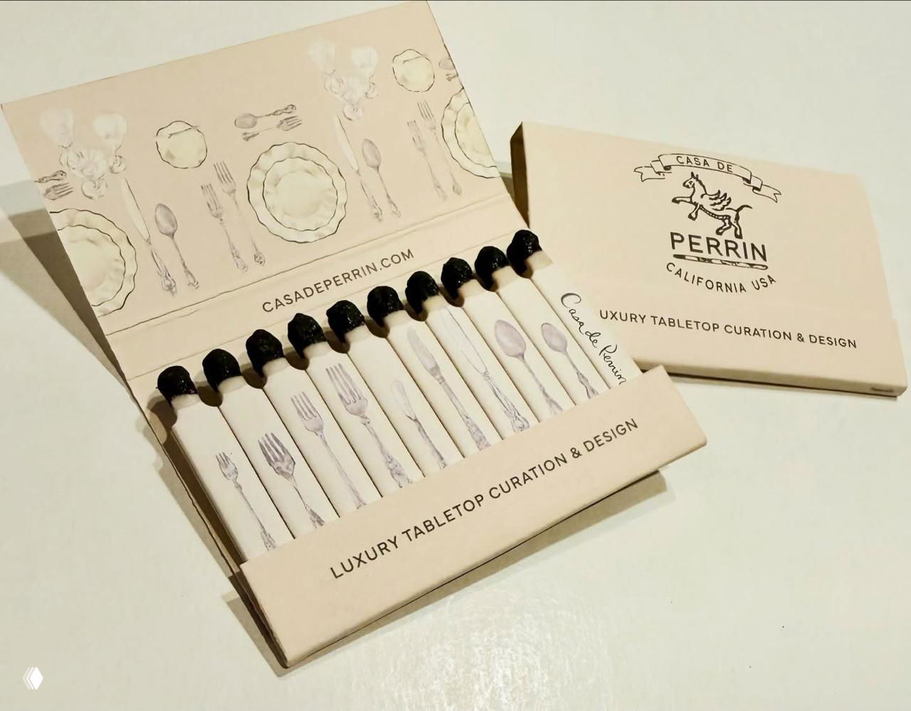 Бежевая коробка 'Casa de Perrin' с иллюстрациями столовых приборов: раскрытая упаковка и спички с рисунками вилок, ложек и тарелок.