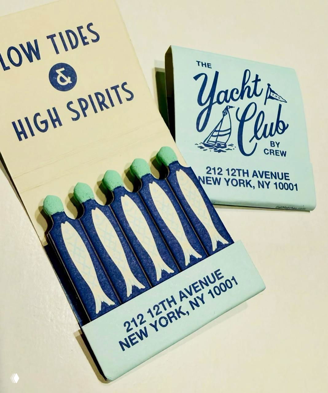 Светло-голубая коробка 'The Yacht Club' с морской темой: раскрытая упаковка и пять спичек в форме рыб с зелёными головками, адрес клуба на поле.