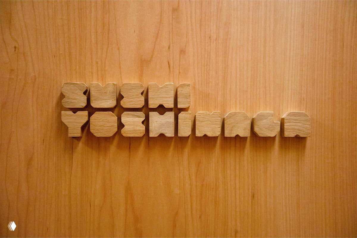 Объёмный деревянный логотип, выточенный и закреплённый на стеновой панели; тёплая текстура дерева контрастирует с минималистичной геометрией знака ресторана