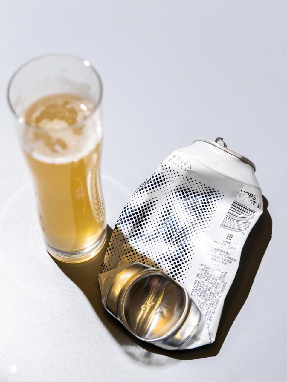 Смятая алюминиевая банка с белой растровой этикеткой рядом со стаканом с пивом: видна текстура, штрихкоды и техническая информация на обратной стороне