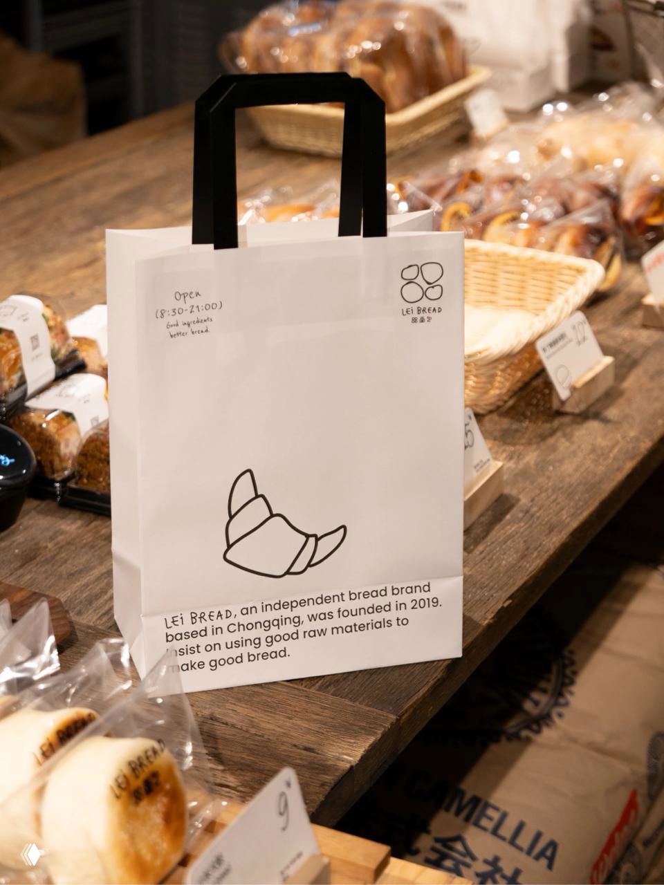 Фотография бумажного пакета Lei Bread на прилавке пекарни: белый пакет с черной ручкой, простая линейная иллюстрация круассана и рукописные надписи.