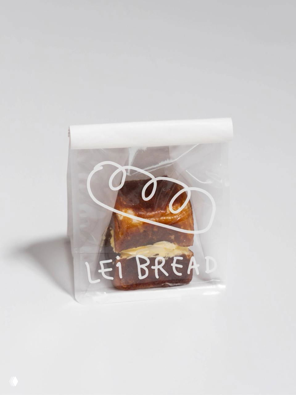Маленькая упаковка с сдобной выпечкой Lei Bread: прозрачный пакет с белой рукописной надписью и стилизованной графикой булочки на светлом фоне.