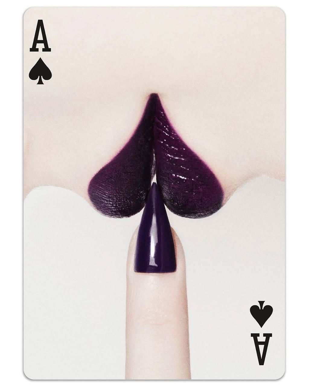 Крупный план карты с фиолетовыми губами и ногтем, оформленными как масть, минималистичный белый фон акцентирует фотографическую постановку.