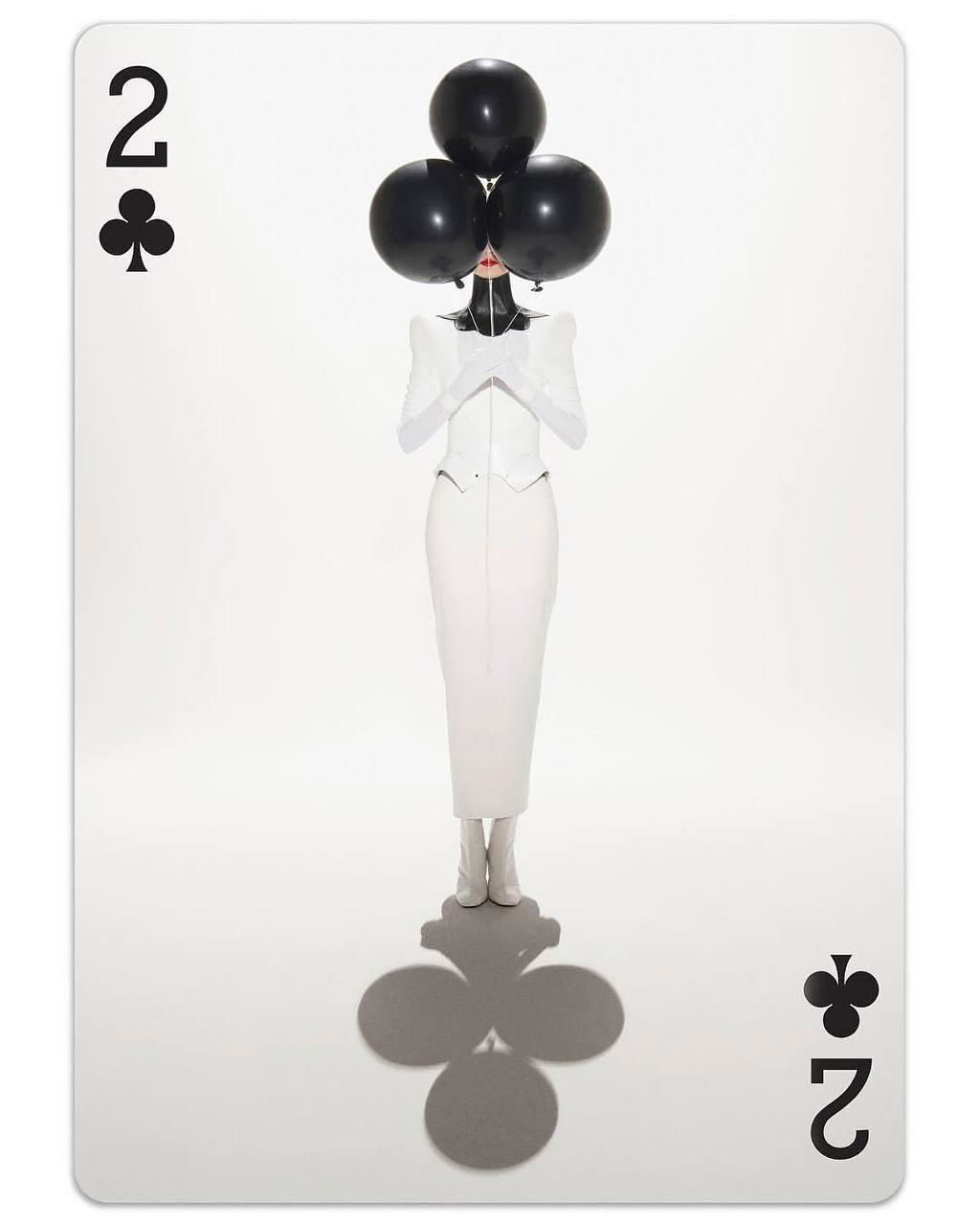 Портретная карта с фигурой в белом и тремя черными шарами, создающими форму масти в тени на полу — игра силуэтов и объектов.