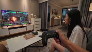 Домашняя игровая сцена: девушка держит джойстик и играет в файтинг на большом телевизоре; гостиная с предметами мебели и уютной обстановкой на заднем плане.