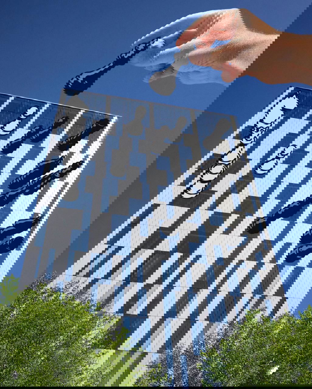 Крупная рука помещает вырезанную шахматную фигуру над фасадом высотного здания, создавая иллюзию шахматной доски на фасаде.