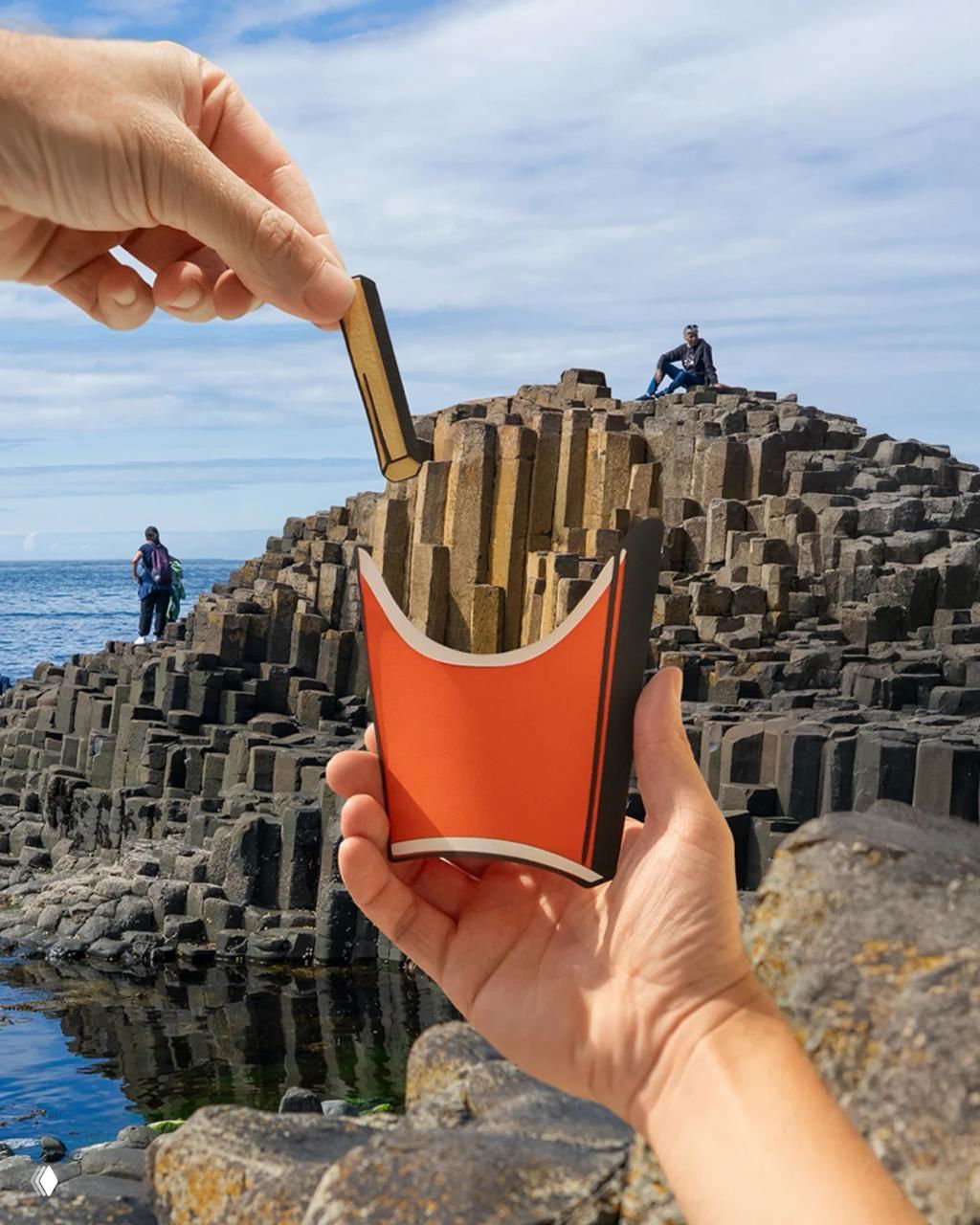 Две руки держат бумажную конструкцию перед базальтовыми колоннами, создавая игру масштаба и пластики форм на причале.