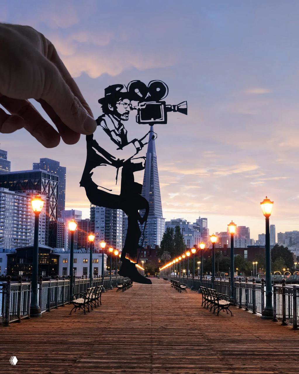 Чёрно‑белая бумажная фигура режиссера с камерой на фоне набережной и небоскрёба, рука автора в кадре создает театральный эффект.