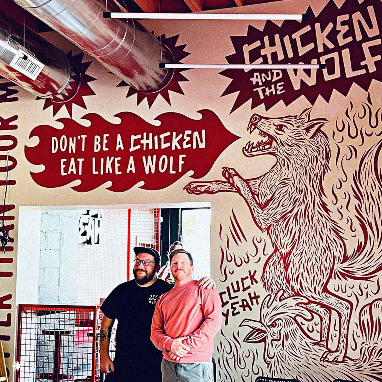 Интерьерное фото: настенная роспись с волком и слоганом «Don't be a chicken, eat like a wolf», авторы проекта на фоне мурала — контекст использования оформления.