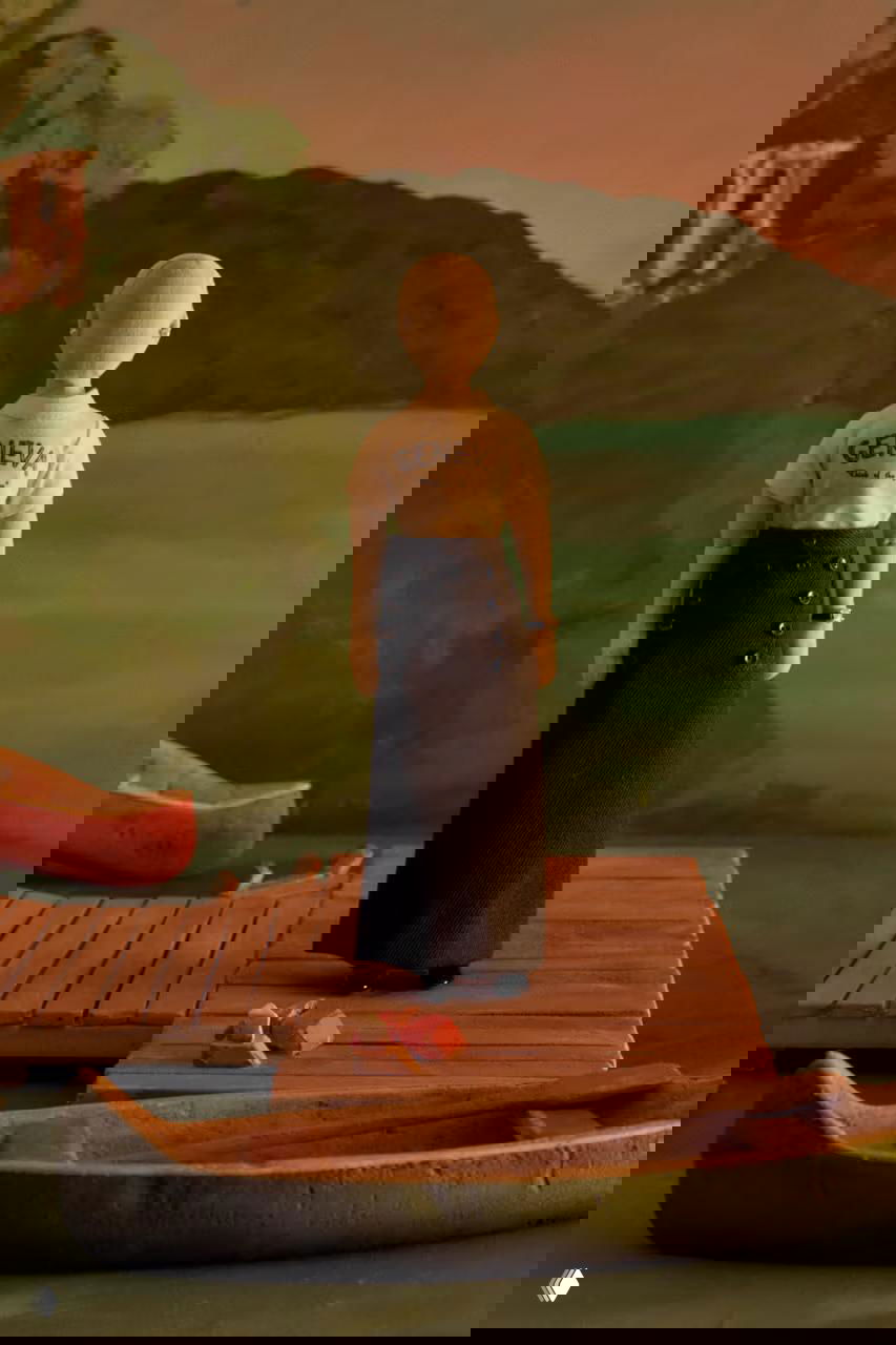 Кукла спиной в футболке с надписью "GENEVA" на пирсе, вокруг миниатюрные лодки и живописный фон в тёплой гамме.