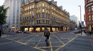 Сцена Genie 3: велосипедист едет по перекрёстку в европейском городе, городской фон и архитектура, фотографическая стилизация