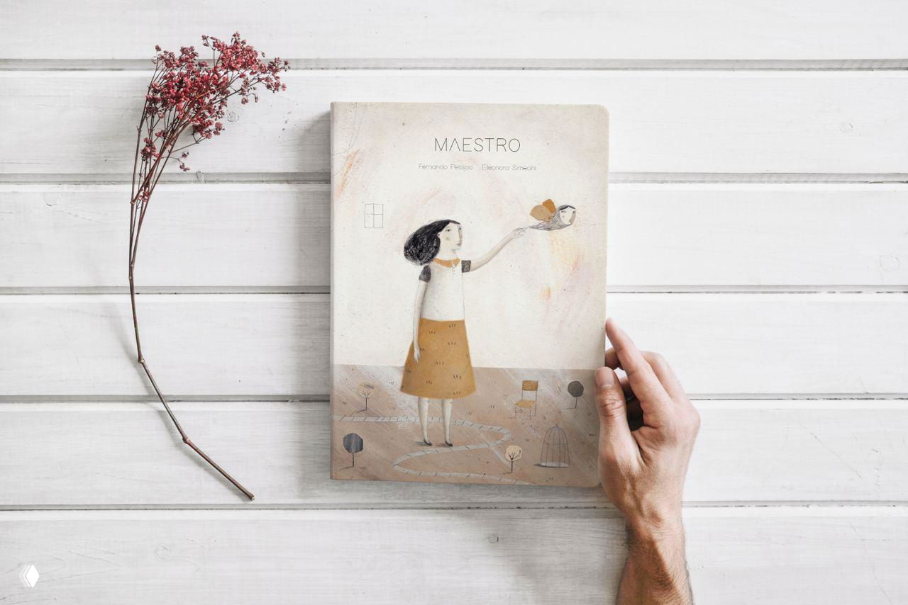 Фотография обложки книги Maestro на деревянной поверхности, рядом сухой цветок и рука держит издание — демонстрация физического макета проекта иллюстраций.