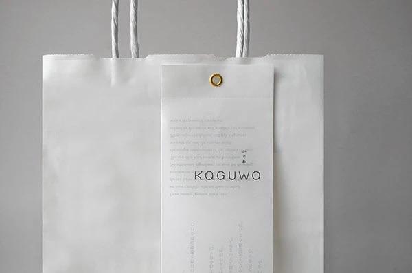 Белый бумажный пакет с подвешенной длинной биркой, на которой тонкой типографикой указан бренд Kaguwa; фон нейтральный серый