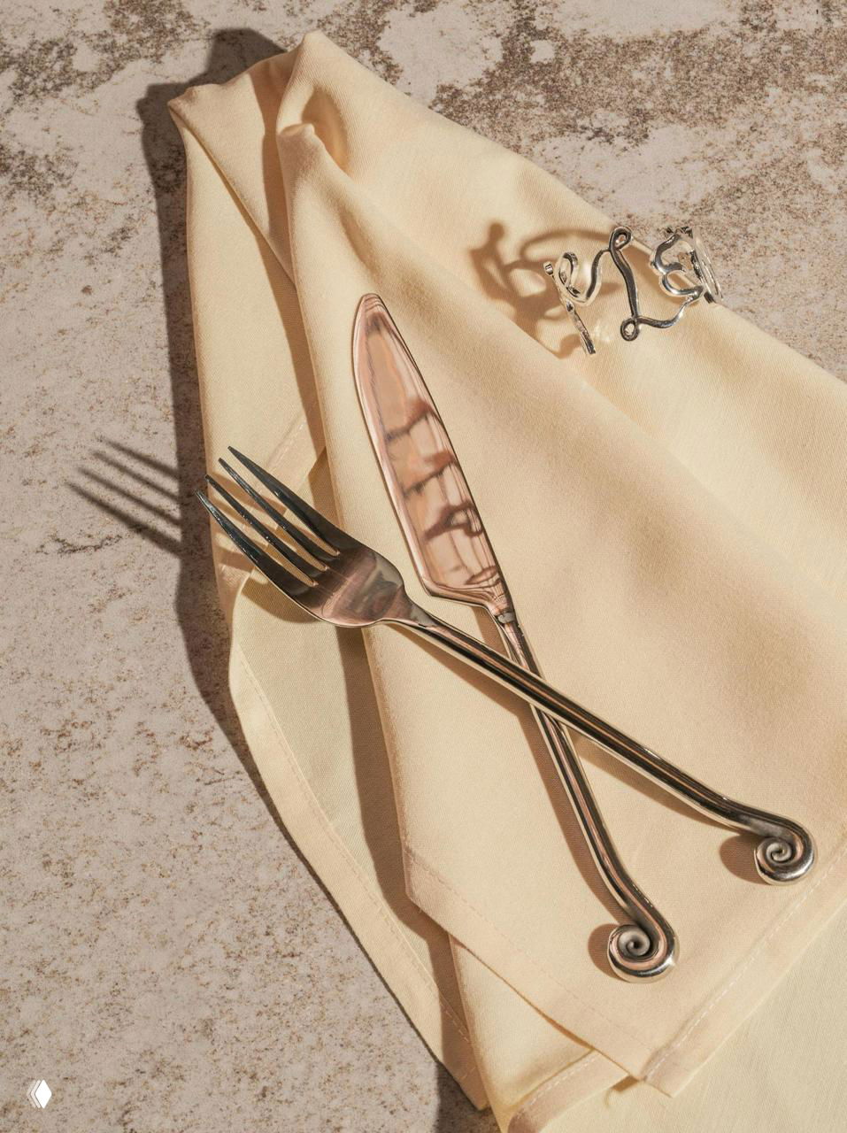 Набор столовых приборов со завитками и кольцо‑салфетник на кремовой салфетке; натюрморт демонстрирует металлические завитки, напоминающие раковину.
