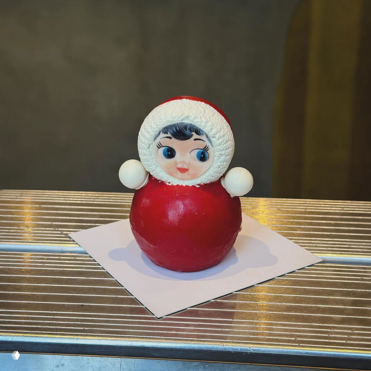 Круглый красный десерт в форме матрёшки с лицом из мастики, установленный на белой подложке; контрастная фактура и детальная отделка видны на фото.