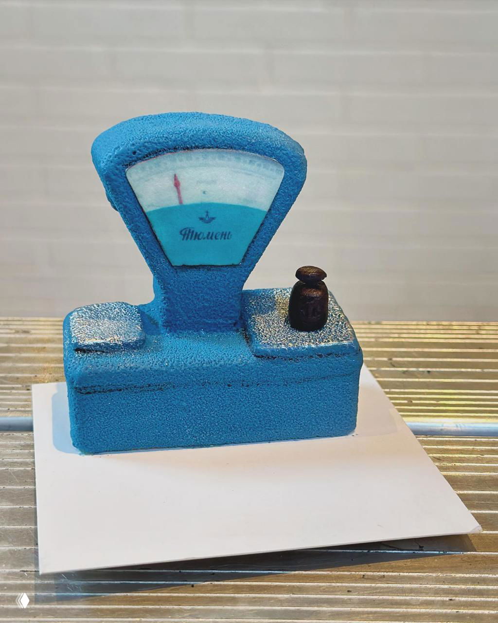 Голубой десерт в виде старинных весов с имитацией циферблата и маленьким грузиком, снят на нейтральном фоне крупным планом.