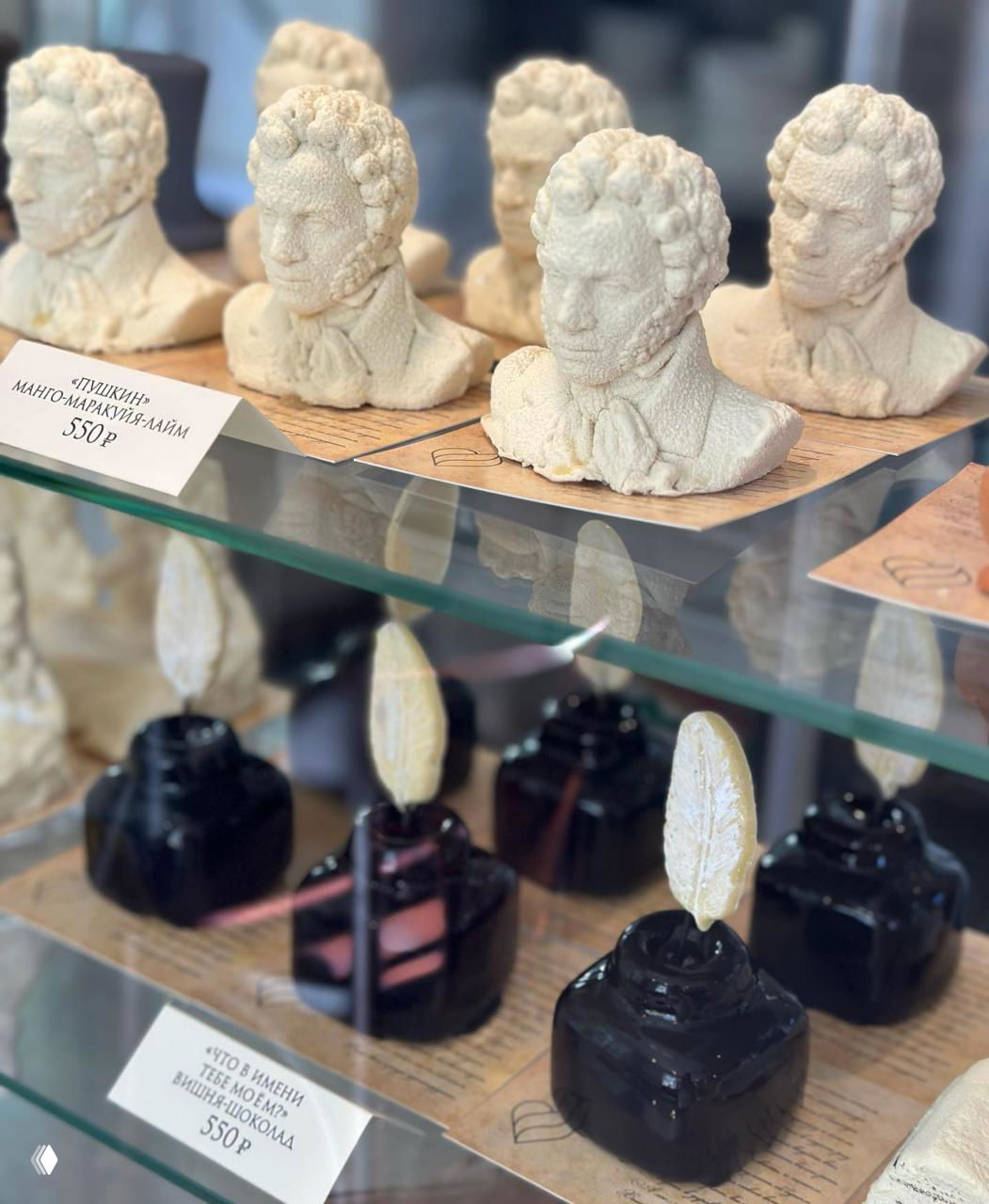 Белые объёмные мини‑бюсты писателя из кондитерской массы на витринной полке, рядом видны тёмные глянцевые пирожные — ассортимент кондитерской.