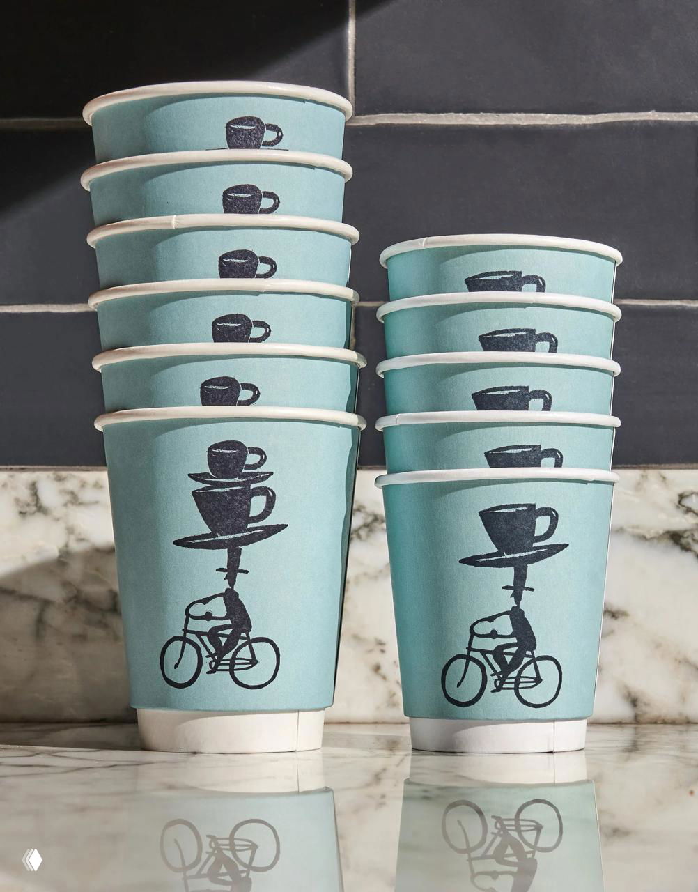 Стопка светло-бирюзовых бумажных стаканов с ручными иллюстрациями чашек и велосипедов — фирменная упаковка кафе на мраморном столе.