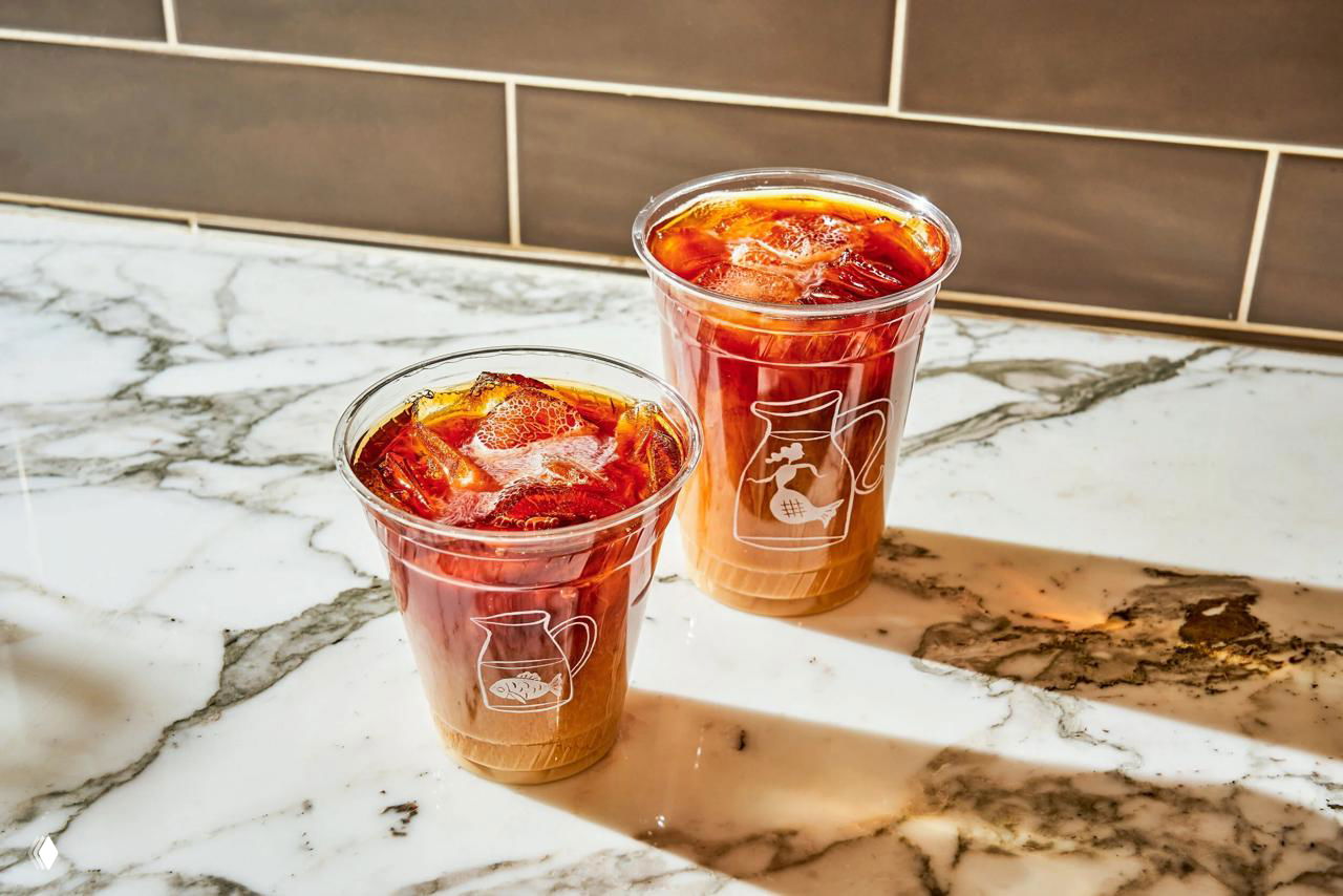 Два пластиковых стакана со светлыми айсед-кофе и белыми графическими логотипами кувшина — фотография напитков с отражениями на мраморе.