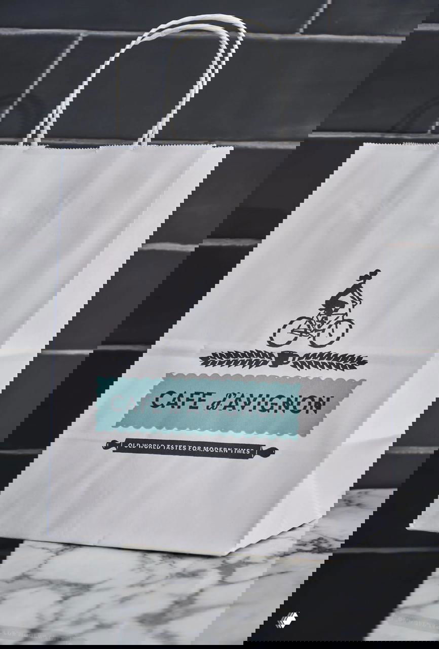 Бумажный пакет с бирюзовой печатью «CAFE d’AVIGNON» и силуэтом велосипедиста — брендирование упаковки для выноса на фоне плитки.