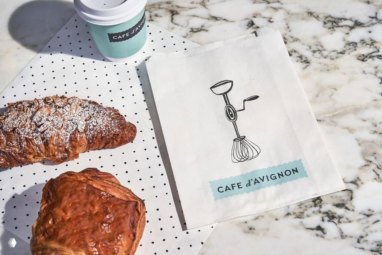 Круассаны и бумажный пакет с иллюстрацией венчика и надписью CAFE d’AVIGNON на мраморном столе — съёмка выпечки и упаковки.