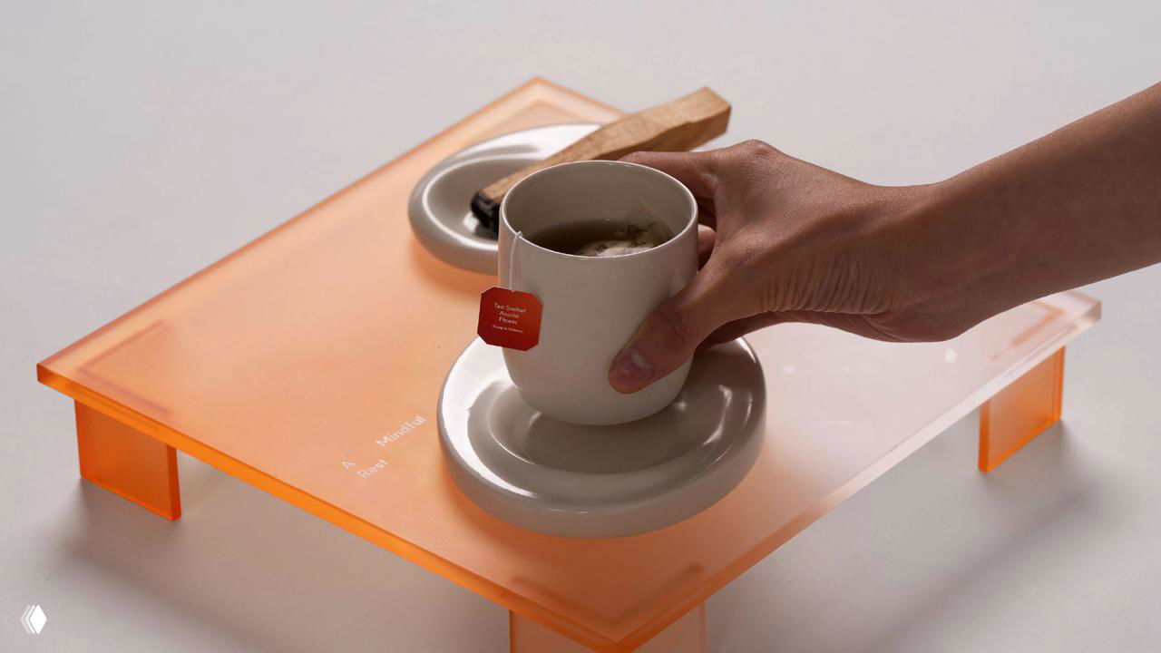 Рука ставит чашку с яркой чайной биркой на прозрачный оранжевый поднос; видна композиция, благовония и элементы сервировки в наборе.