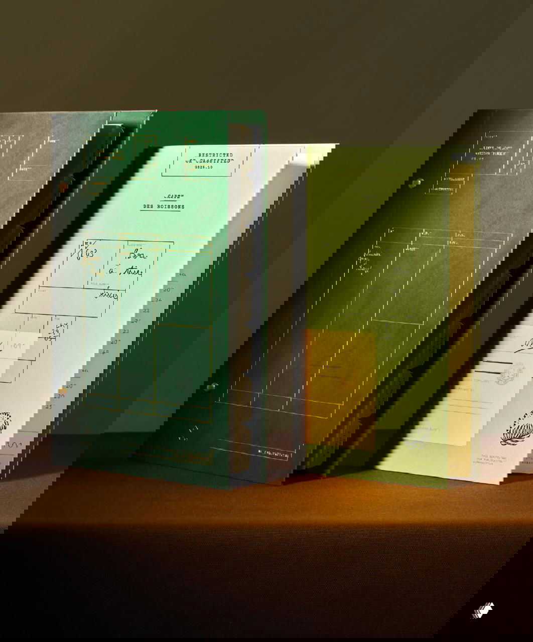 Несколько папок и меню в зелёно-жёлтой гамме, стоящие на столе: видны печать, тиснение и вкладные карточки — комплексная система печатных материалов для ресторана.